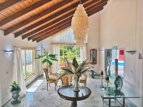 A spacious room with a high ceiling, a large chandelier, a dining area with chairs, a table, and a decorative mirror.