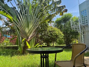 A garden with a table and chair, surrounded by lush greenery and a white building with a spiral staircase.