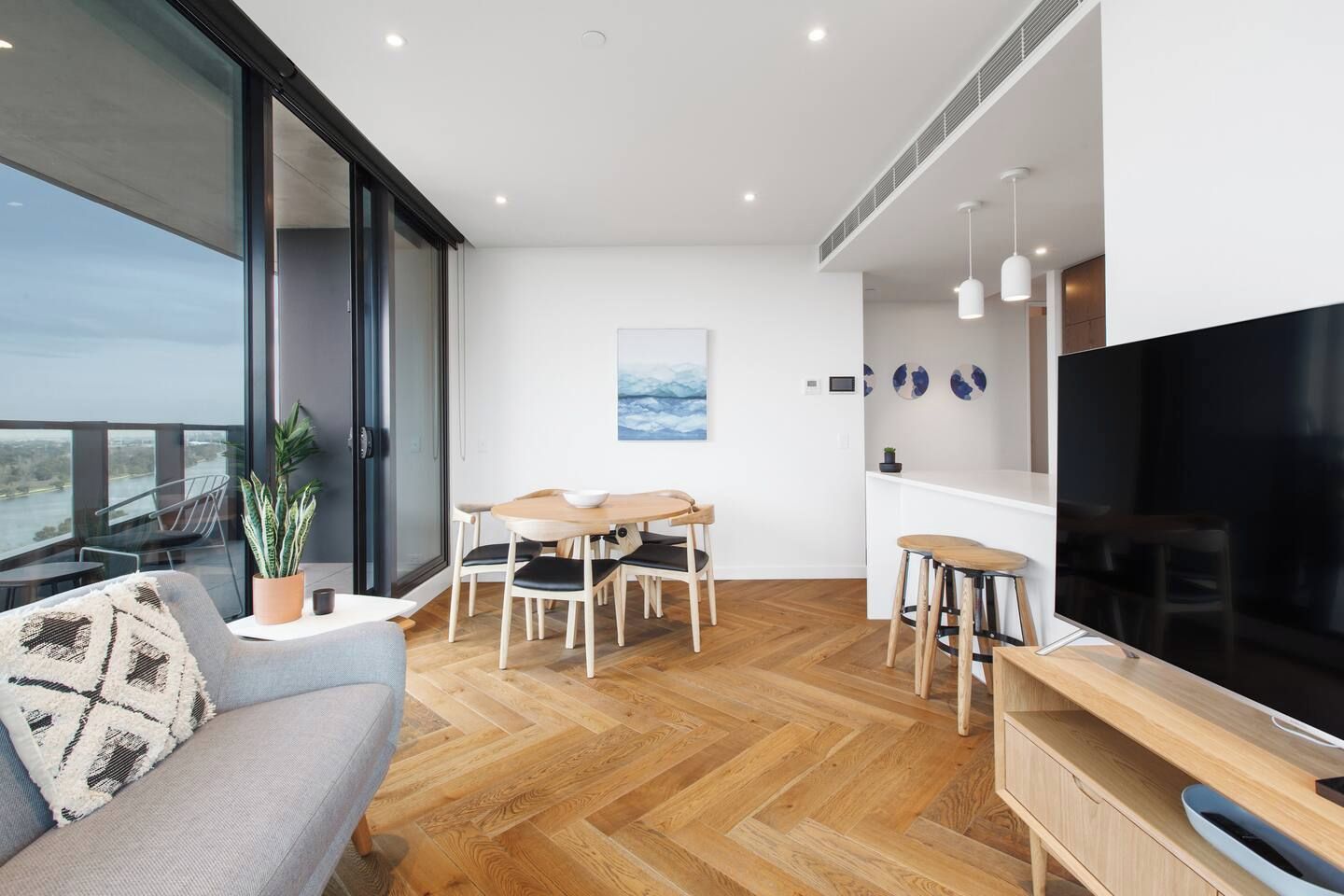 A modern living room with a large TV, wooden flooring, and a balcony with a view.