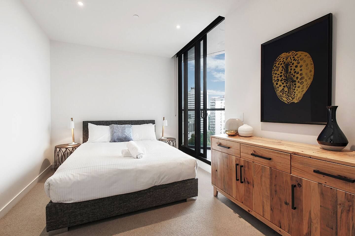 A modern bedroom with a large bed, a wooden dresser, a city view, and a leopard print artwork.