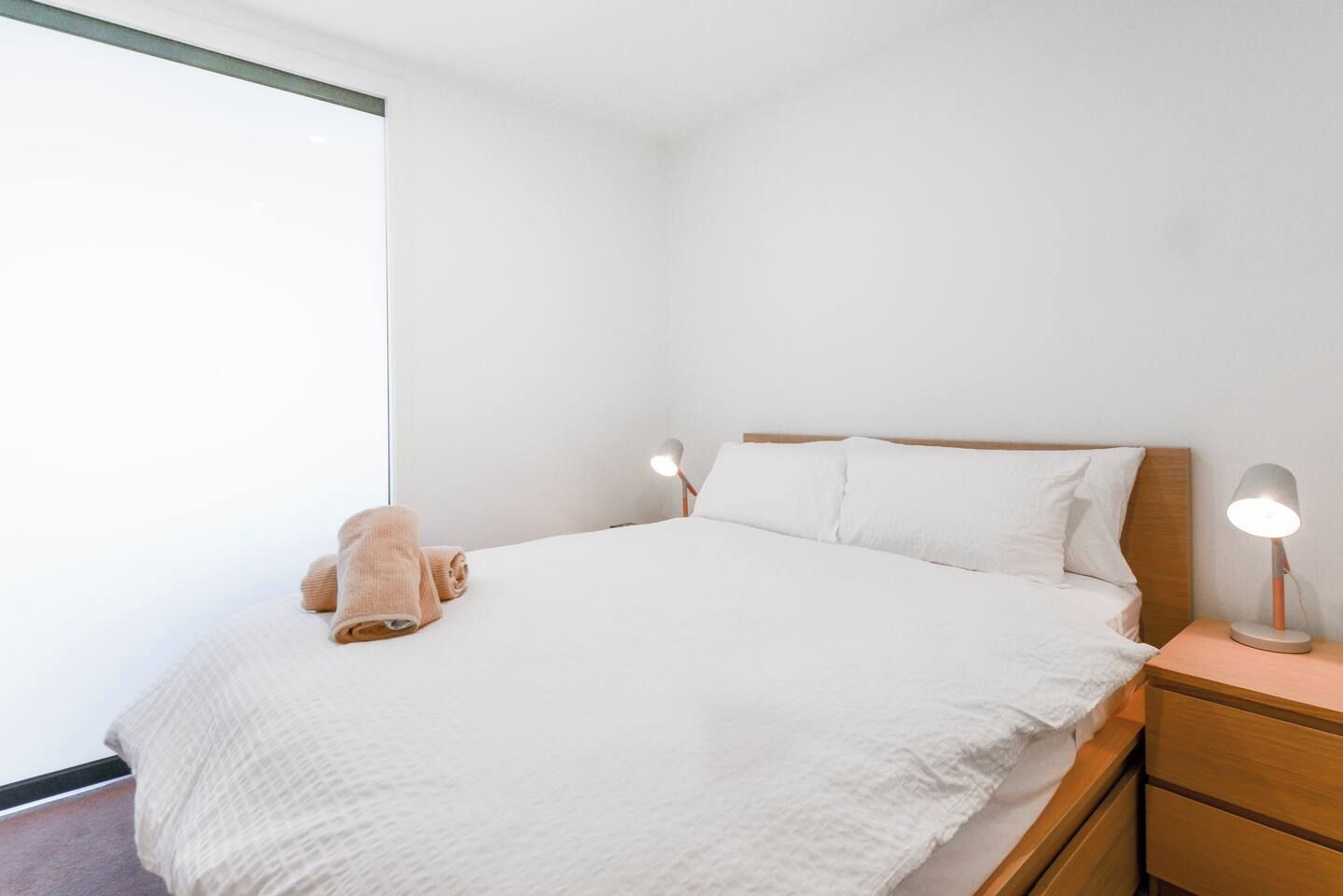 A neatly made bed with white linens and a single rolled-up towel, a bedside table with a lamp, and a window with a view.