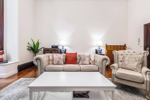 A living room with a sofa, armchair, and coffee table. There are two lamps, a plant, and a wooden cabinet. - Elwood Manor A Restored Art Deco Gem (Elwood)
