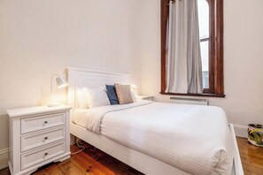 A neatly made bed with white bedding, a bedside table with a lamp, and a window with curtains. - Elwood Manor A Restored Art Deco Gem (Elwood)