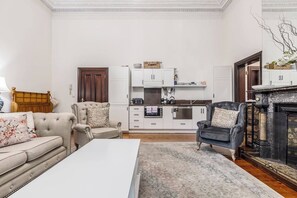 Ein Wohnzimmer mit einer Couch, Sesseln, einem Couchtisch und einem Kamin. Im Hintergrund ist eine Küche mit weißen Schränken und einem Steinkamin zu sehen.