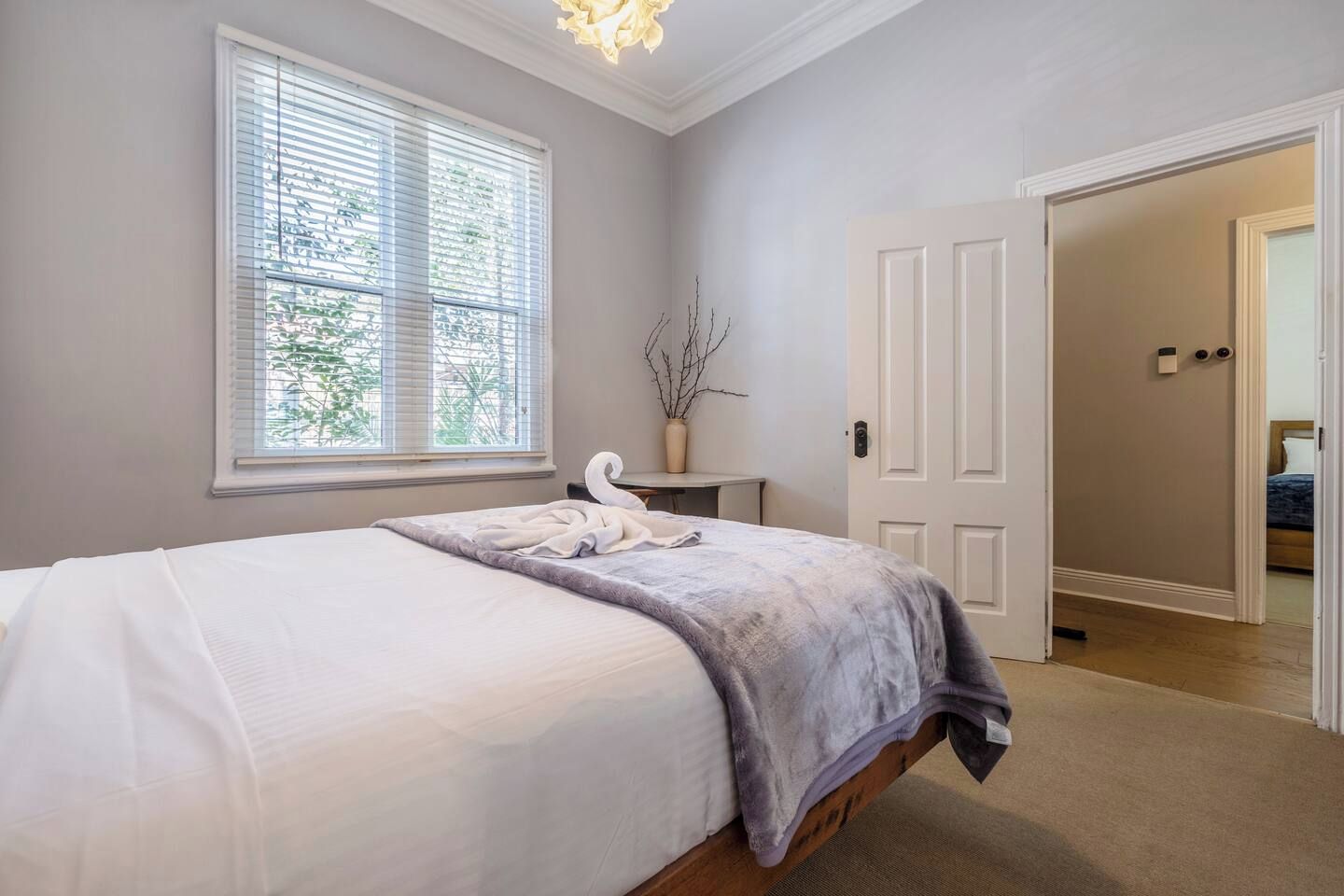 A bedroom with a bed, a window with blinds, a vase with branches, and a door leading to another room.