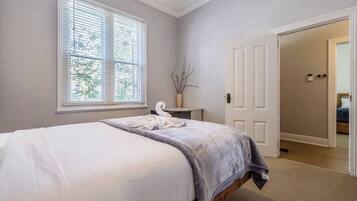 A bedroom with a bed, a window with blinds, a vase with branches, and a door leading to another room.