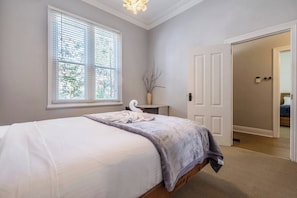 A bedroom with a bed, a window with blinds, a vase with branches, and a door leading to another room. - Classic Comforts Moments to Flemington Racecourse (Flemington)