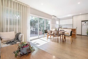 Ein modernes Wohnzimmer mit einer Couch, einem Esstisch mit Stühlen, einer Küchenzeile mit Schränken und einer Glastür, die auf eine Terrasse führt.