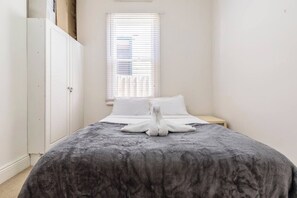 Ein Schlafzimmer mit einem Bett, weißen Kissen, einer grauen Decke, einem weißen Handtuch, einem Nachttisch aus Holz und einem Fenster mit Jalousien.