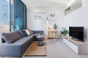 A modern living room with a grey sofa, a small wooden desk, a TV, and a large window. - Dazzling Views from Contemporary High Rise Living (Melbourne)