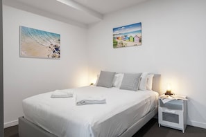 A neatly made bed with pillows and a folded towel, a bedside table with a lamp and a book, and two wall-mounted paintings of beach scenes. - Skyline Panorama in Melbourne CBD with Parking (Melbourne)