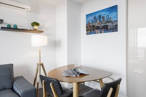 A modern hotel room with a round table, two chairs, a lamp, a cityscape painting, and a small shelf with decorative items. - Skyline Panorama in Melbourne CBD with Parking (Melbourne)