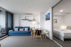 A modern room with a blue sofa, a dining table, and a bed. There are wall-mounted shelves with decorative items. - Skyline Panorama in Melbourne CBD with Parking (Melbourne)