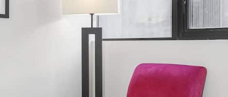 A room with a bright window, a floor lamp, a pink chair, and a white side table.