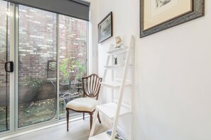Habitació amb porta corredissa de vidre que dona a una façana de maó, una cadira de fusta, una escala blanca i una estant amb objectes decoratius.