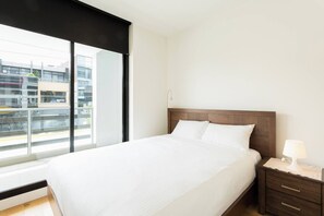 A hotel room with a large window, a bed with white linens, a wooden headboard, a nightstand with a lamp, and a view of buildings outside. - Urban Stay in Trendy Windsor close to Chapel St (Windsor)