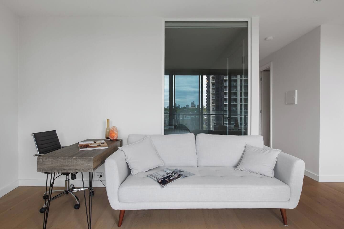 Een moderne woonkamer met een witte bank, een houten salontafel en een zwarte bureaustoel. Er is een groot raam met uitzicht op de stad.