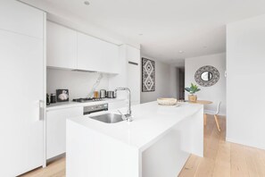 A modern kitchen with white cabinetry, a stainless steel sink, and a built-in stove. There is a small dining area with a round table and a chair. - Executive Resort Style Living at Leafy Albert Park (Melbourne)