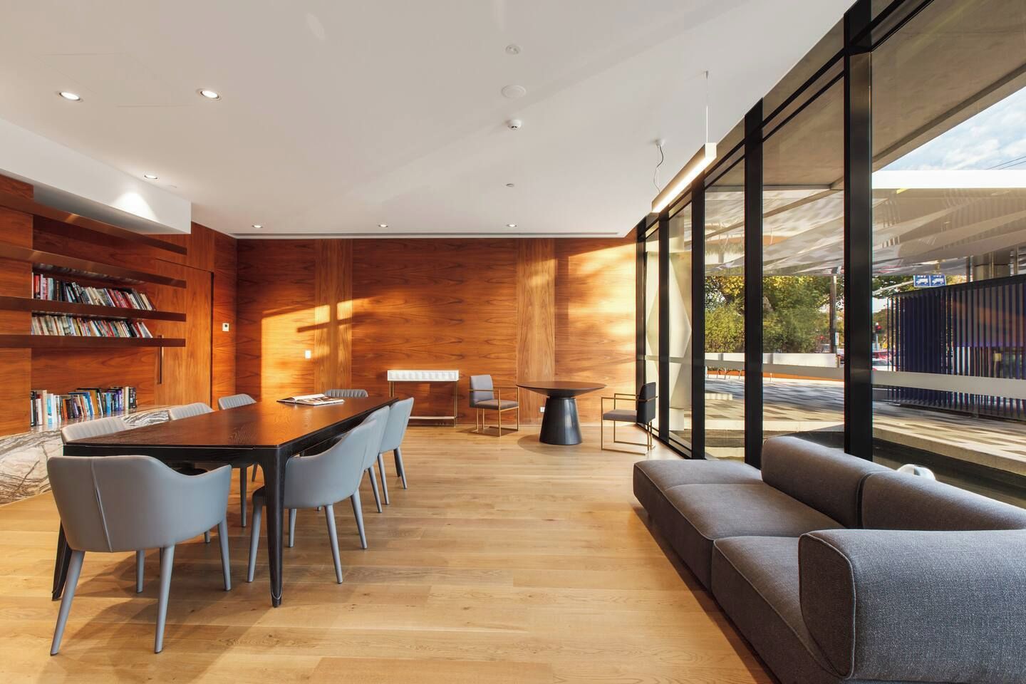 A modern interior with a wooden wall, a dining area with a long table and chairs, a sofa, and large windows offering a view of the outside.