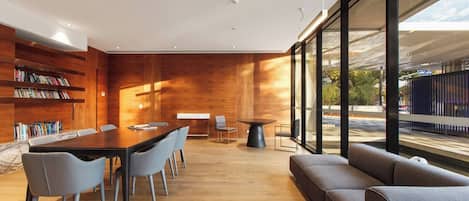 A modern interior with a wooden wall, a dining area with a long table and chairs, a sofa, and large windows offering a view of the outside.