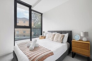 A modern bedroom with a large window, a bed with white linens, a grey upholstered headboard, a bedside table with a lamp, and a small clock. - Executive Resort Style Living at Leafy Albert Park (Melbourne)