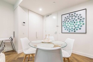 A modern dining area with a glass table surrounded by white chairs, a framed abstract art piece on the wall, and a white cabinet with a door. - Parque Canopy Lakeside Living with Infinity Pool (Melbourne)