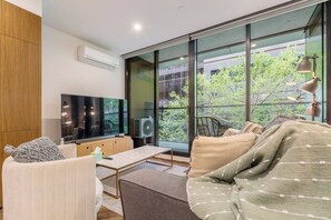 A modern living room with a flat-screen TV, comfortable seating, and large windows offering a view of greenery. - Parque Canopy Lakeside Living with Infinity Pool (Melbourne)