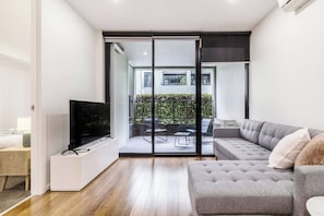 A modern living room with a grey sofa, a flat-screen TV, and a sliding glass door leading to an outdoor patio with a table and chairs. - Nottville Poolside Coastal Cosmopolitan off Bay St (Port Melbourne)