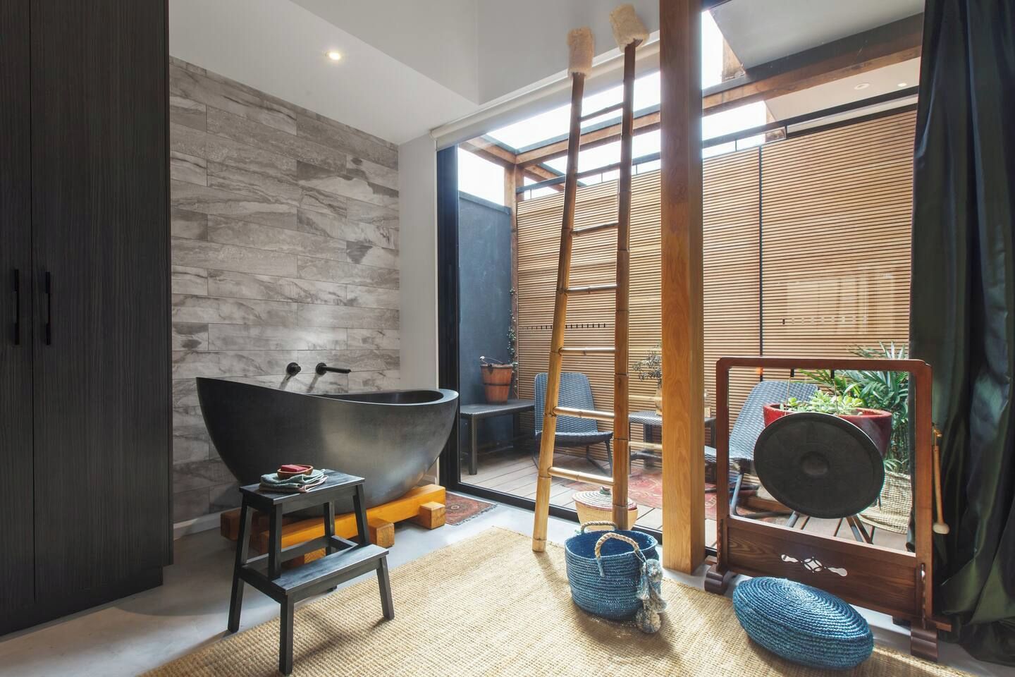 A modern bathroom with a freestanding bathtub, a wooden ladder, and a view of an outdoor seating area.