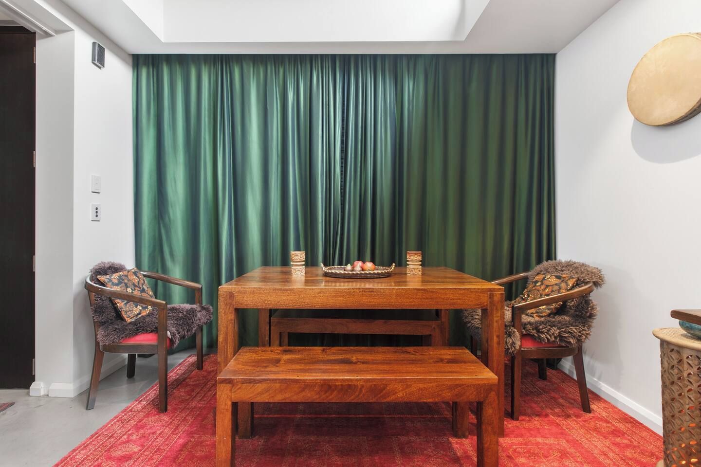 A dining area with a wooden table and benches, a green curtain backdrop, and a red patterned rug.