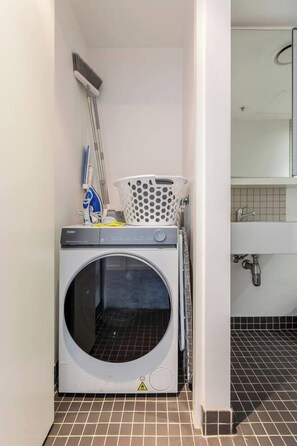 A compact laundry area with a front-loading washing machine, a mop, and a laundry basket. - Mordyn at Victoria Brewery Parkside Escape w Pool (East Melbourne)