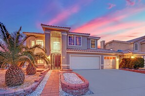 A two-story house with a garage, a driveway, and a landscaped front yard with palm trees and a brick pathway. - Luxury Vegas Escape 4BR, Pool/Spa, Game Room & BBQ (Las Vegas)