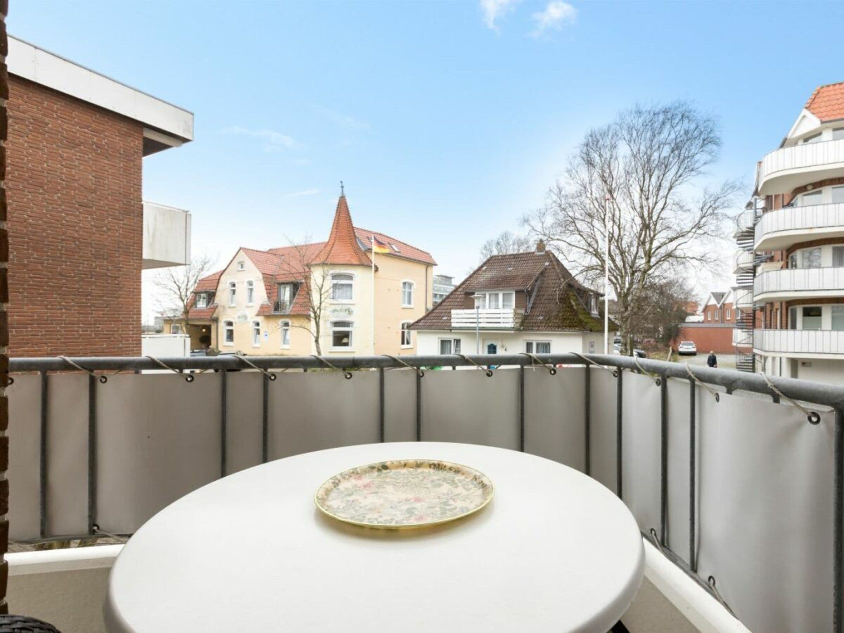 A balcony with a round table and a plate, overlooking a row of houses with a prominent building with a red roof.