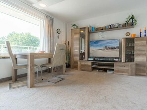 A modern living room with a wooden dining table, a comfortable chair, a flat-screen TV, and a bookshelf. - Holiday Apartment 19 (St. Peter-Ording)