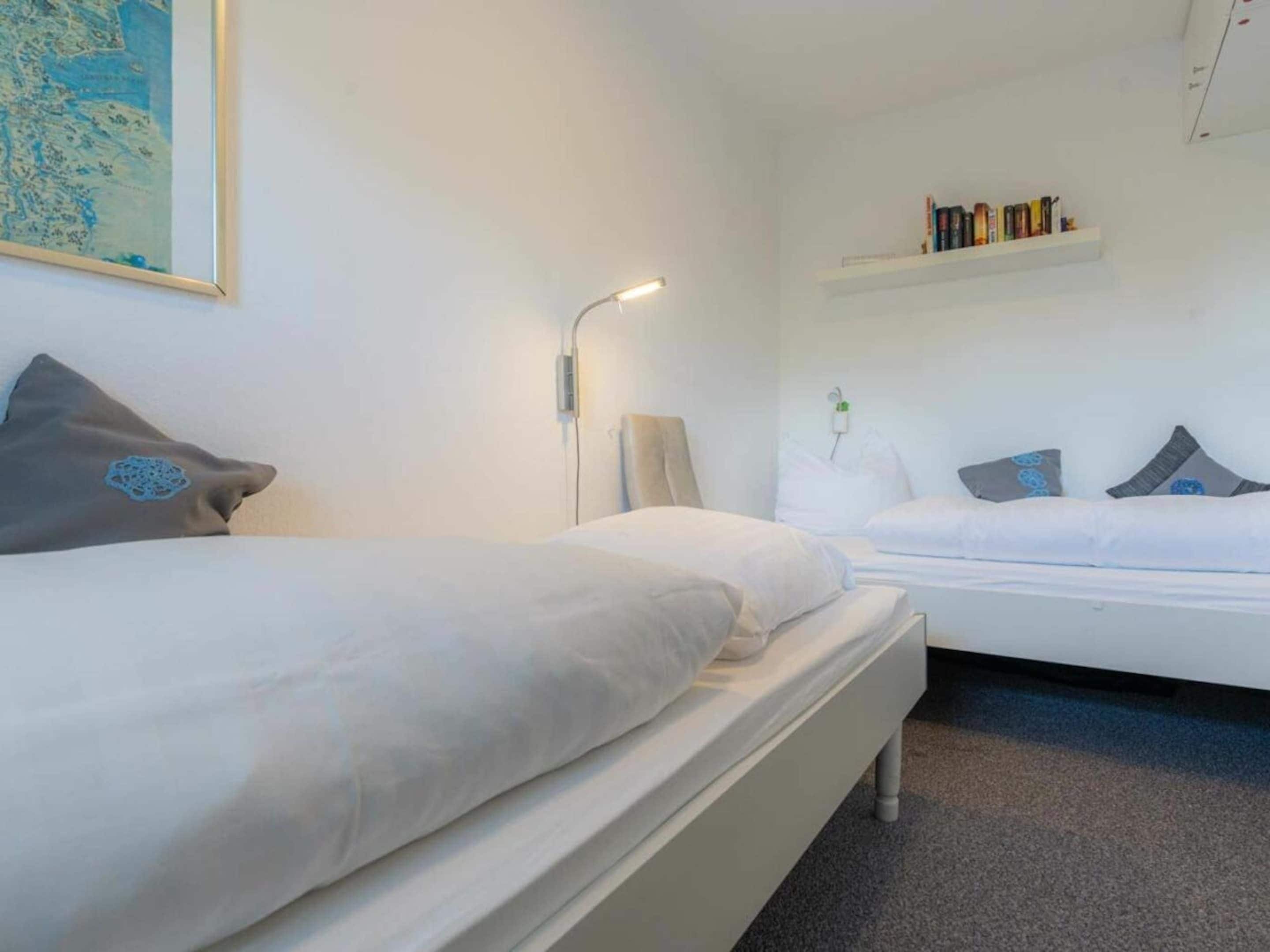 A hotel room with two beds, a bedside table, a lamp, a wall-mounted shelf with books, and a framed map.