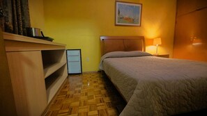 Una habitación pequeña y funcional con cama, mesita de noche con lámpara, armario de madera y un electrodoméstico blanco pequeño.