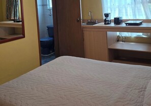 Un dormitorio con una cama, un escritorio de madera con una computadora, una cafetera y una ventana con cortinas.
