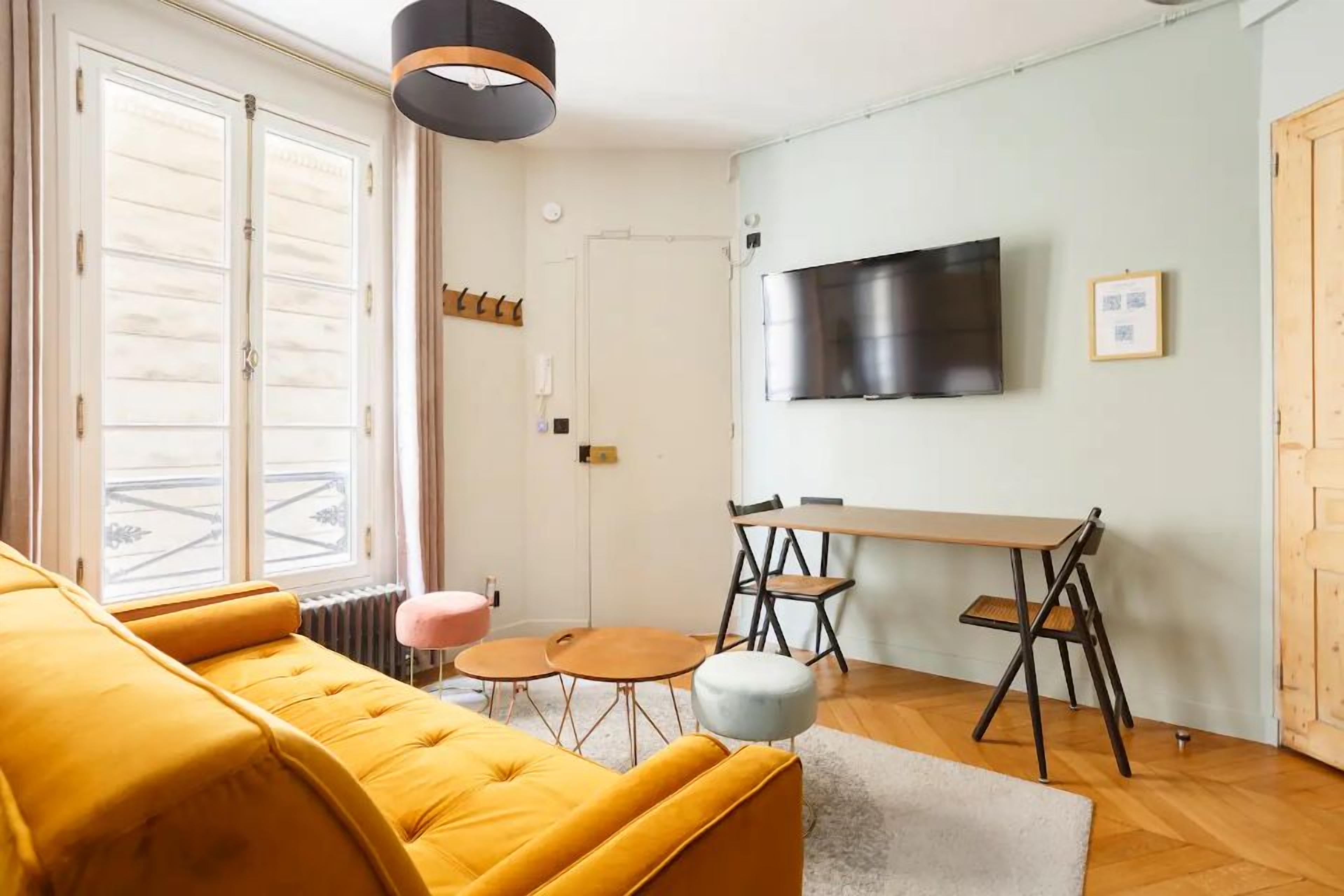 Una sala de estar moderna con un sofá amarillo, una mesa de comedor con sillas, un televisor montado en la pared y un ventanal con vistas.