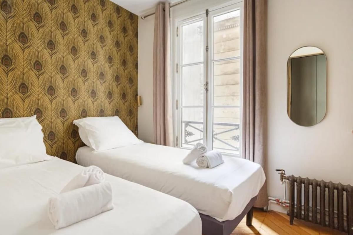 A hotel room with two beds, a window with curtains, a radiator, and a mirror on the wall.