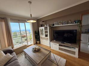 Una sala de estar con televisor grande, sofá, mesa de centro con adornos, aparador con fotos enmarcadas y balcón con vista a edificios y follaje.