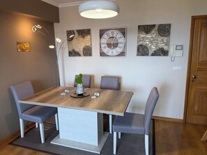 A dining area with a wooden table, grey upholstered chairs, a vase with a plant, and wall art including a clock and abstract paintings. - Your peaceful home in Funchal—enjoy the garden and the sea (funchal)