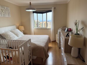 A bedroom with a bed, a crib, a dresser, a lamp, and a window with a view of buildings. - Your peaceful home in Funchal—enjoy the garden and the sea (funchal)