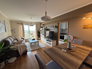 A modern living room with a sofa, a dining table, and a kitchen area with cabinets and a stove. - Your peaceful home in Funchal—enjoy the garden and the sea (funchal)