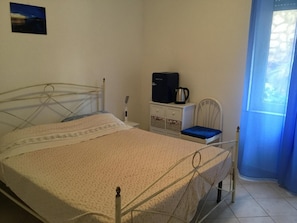 Una cama individual con estructura metálica, una mesita de noche con hervidor, una silla y un mueble auxiliar pequeño.