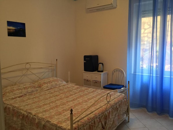 Un dormitorio con una cama, un mueble blanco pequeño, un electrodoméstico negro y una ventana con cortinas azules.
