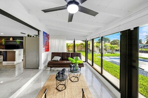 A modern living room with a large window, a ceiling fan, and a view of a landscaped garden. - Wilton Manors Waterfront Villa w/ Gorgeous Pool (Wilton Manors)