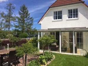 A two-story house with a glass conservatory, a patio with a table and chairs, and a well-maintained garden. - Holiday Home in Putbus With Swimming Pool (Putbus)