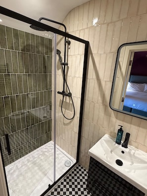 Casa de banho com chuveiro em cabine de vidro, lavatório branco e chão xadrez preto, com um espelho a refletir uma cama.