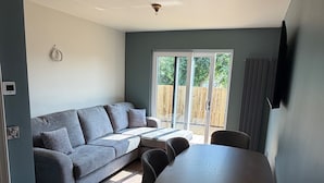 A modern living room with a grey sofa, a dining table with chairs, and a sliding glass door leading to a wooden deck. - House Sleeps 8 With Parking and Balcony (Clones)