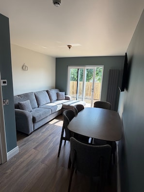 Sala de estar moderna com sofá cinza, mesa de jantar com cadeiras e uma porta de vidro deslizante para um deck de madeira.